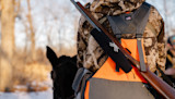 Hunter in camouflage with orange pack, rifle slung on his back, horse head beside him