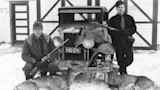 Two hunters with rifles by vintage truck and four deer carcasses in snow, sign reads OFFICE