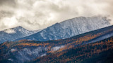 Snow-dusted mountain peaks above layered forested ridges with golden trees and cloudy sky
