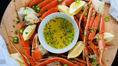 Bowl of melted herb-and-chili butter on wooden board surrounded by crab legs, lemon wedges, sliced jalapeño