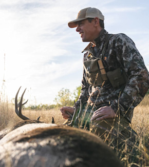 Hunter kneeling beside a buck with antlers in grassy field, wearing camo jacket and cap