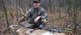 Hunter kneeling behind large buck, holding antlers; compound bow rests on deer in woods