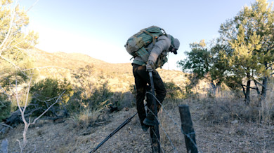 Hunter climbing barbed-wire fence with large backpack and tripod in scrubby hills
