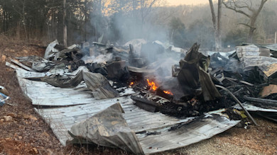 Smoldering cabin ruins with twisted metal roofing, smoke and small flames in wooded clearing