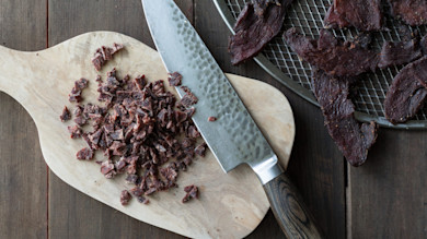 Chopped venison jerky on a wooden cutting board with chef's knife and jerky on wire rack