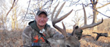 Man kneeling behind large buck holding antlers in leafless woods, Nikon camera strap visible