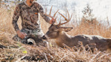 Hunter kneeling in dry grass holding the antlers of a large whitetail buck