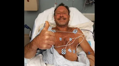 Man in hospital bed giving thumbs-up, chest fitted with ECG electrodes