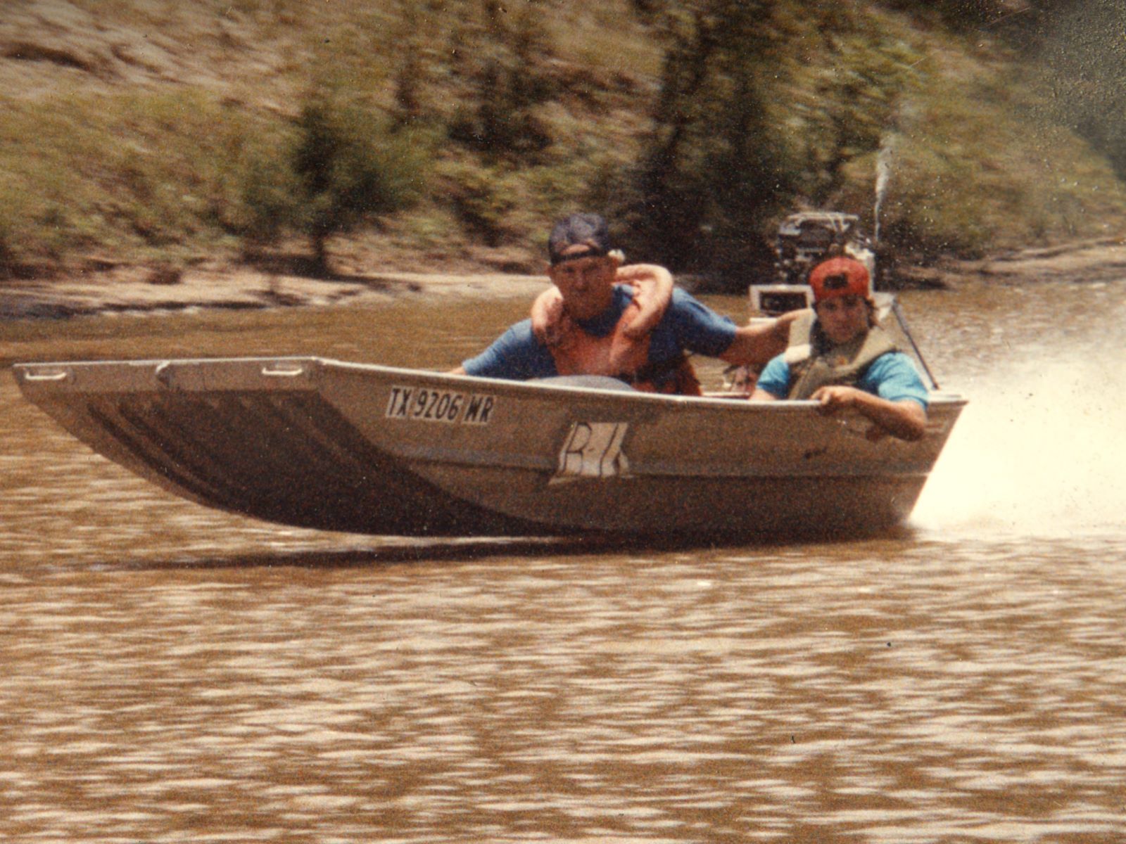 Two men in life jackets speeding an aluminum boat on a brown river; registration TX 9206 NR