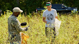 Two men in tall field; one in camouflage handling plants, other wearing gray shirt reading 'I WORK FOR WILDLIFE'