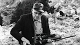 Seated man holding a scoped rifle on a rocky hillside, black-and-white