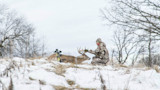 Hunter kneeling and touching buck's antlers, bow resting on deer, snow-covered field