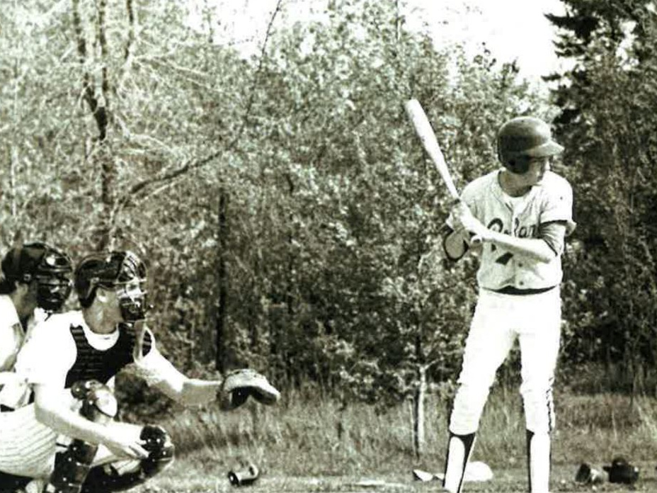 Batter at plate holding bat; catcher crouched with mitt and mask; trees behind.