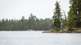 Hunter standing on rock beside a canoe on a misty lake with pine shoreline