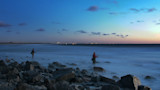 Two anglers wading on rocky shore at dusk, misty surf and distant city lights
