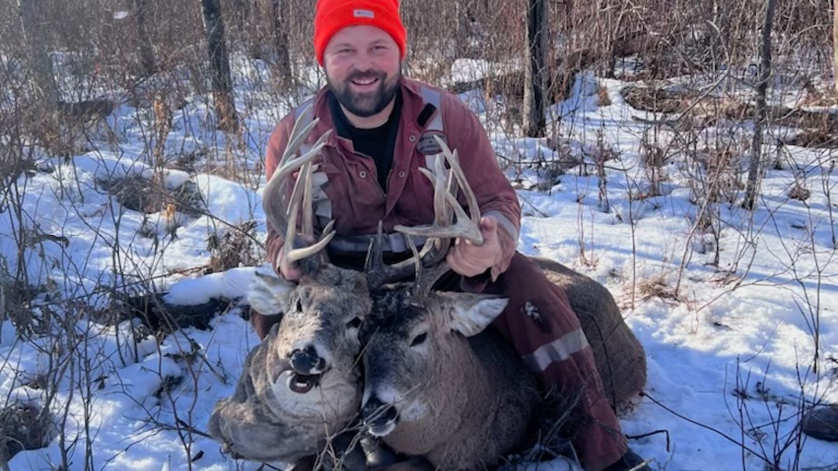 Hunter kneeling in snow holding two deer heads by their antlers, wearing an orange beanie