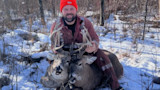 Hunter kneeling in snow holding two deer heads by their antlers, wearing an orange beanie