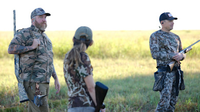 Three hunters in field: bearded man with shotgun and game, woman with ponytail facing away, camo man with shotgun