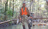 Clay Newcomb standing in a stream carrying a deer and holding a compound bow; cap patch 'BEAR GREASE'