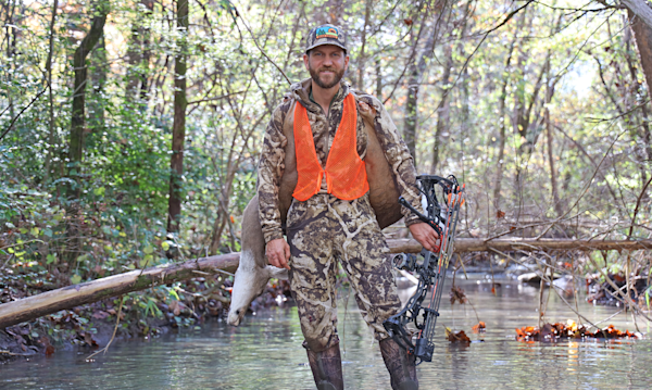 Clay Newcomb standing in a stream carrying a deer and holding a compound bow; cap patch 'BEAR GREASE'