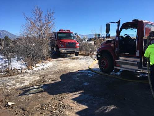 Two red fire trucks on a snowy dirt road; right truck door open with hose; person in neon jacket at right