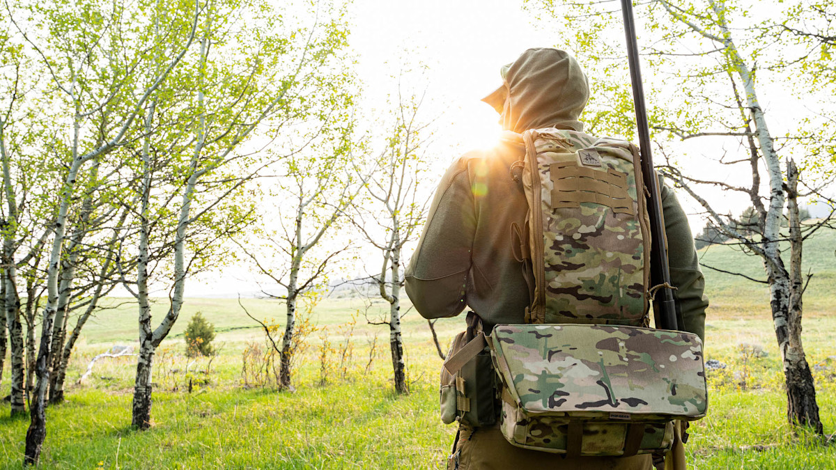 Hunter wearing camo backpack and green hoodie, holding shotgun upright in birch grove with sun flare