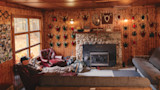 Wood-paneled deer camp with stone fireplace and rows of antler plaques; man reclines in leather chair