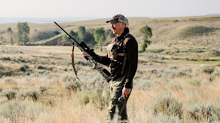 Hunter holding camouflaged rifle in open grassland, wearing cap and chest pack