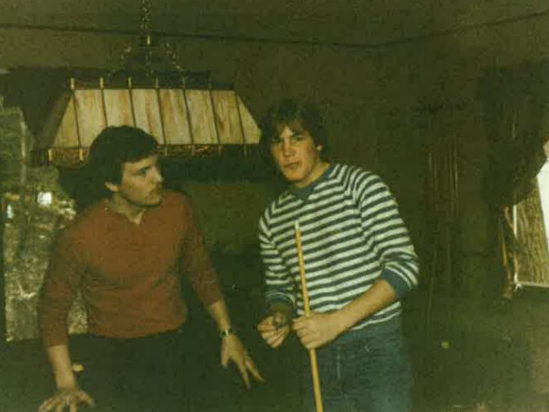 Two young men by a pool table; one wearing blue-striped sweater holding a pool cue