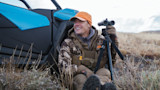 Steven Rinella seated by blue UTV labeled Maverick Trail, holding binoculars on a tripod