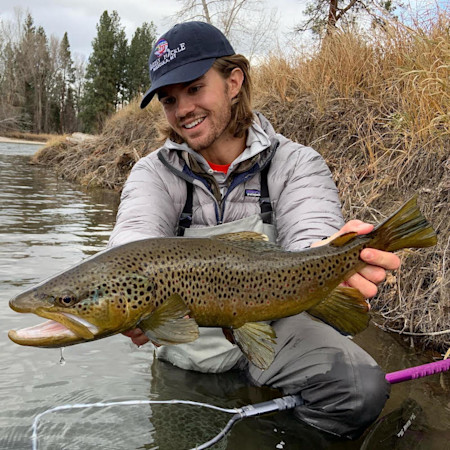 Man kneeling in river holding large brown trout, wearing Patagonia jacket