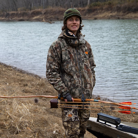 Young hunter by river holding wooden longbow with three orange-fletched arrows, camo.