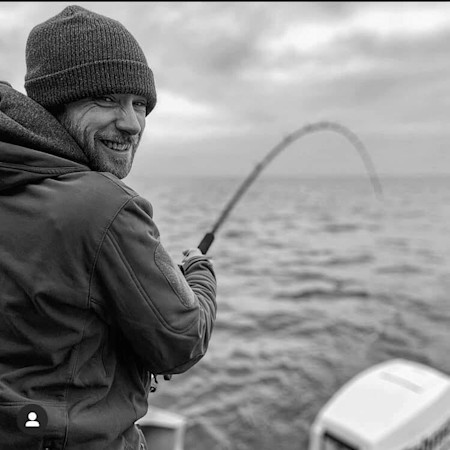 Smiling man fishing from boat with bent rod, wearing knit beanie and hooded jacket