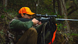 Hunter in orange First Lite cap aiming rifle on backpack in forest