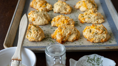 Cheddar drop biscuits on baking sheet sprinkled with herbs, bowl with basting brush nearby