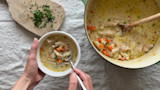 Turkey gnocchi soup in a white bowl held by hands, wooden spoon, pot of soup and chopped herbs