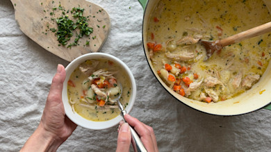 Turkey gnocchi soup in a white bowl held by hands, wooden spoon, pot of soup and chopped herbs