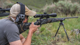 Hunter kneeling, peering through riflescope on camo bolt-action rifle mounted on bipod in grassy field