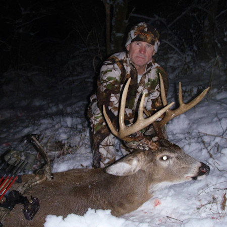 Hunter kneeling beside large buck in snow at night holding antlers; bow nearby