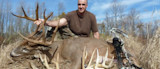 Large buck with tall antlers; man kneels behind it with a compound bow