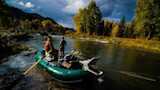 Two anglers in green raft on river with fishing rods, autumn trees and dark storm clouds