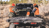 Two hunters in orange vests loading backpacks into a Can-Am UTV bed