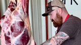 Man with red beard trimming hanging carcass in metal processing shed