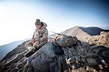 Hunter with rifle and pack seated on rocky mountain ridge with binoculars
