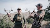 Two hunters holding deer skulls with antlers in dry, hilly shrubland