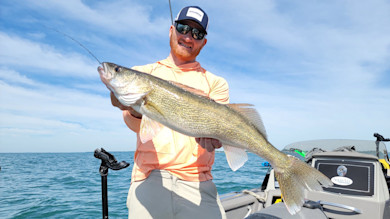 Man holding large walleye on a boat, wearing a cap labeled "FishUSA"
