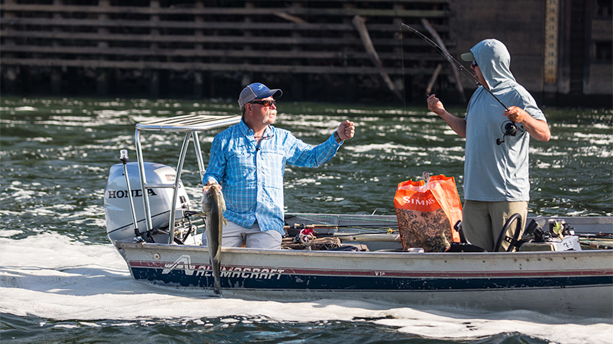 Two men fishing on a boat; left man holds a fish; orange bag reads SIMMS; Honda motor visible