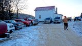 Man in orange beanie and plaid jacket walking on snowy farm road pointing at white farmhouse with parked trucks
