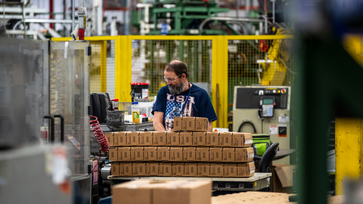 Factory worker at packing table with stacked cardboard boxes, wearing navy shirt reading "USA" and American flag.