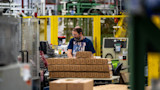 Factory worker at packing table with stacked cardboard boxes, wearing navy shirt reading "USA" and American flag.